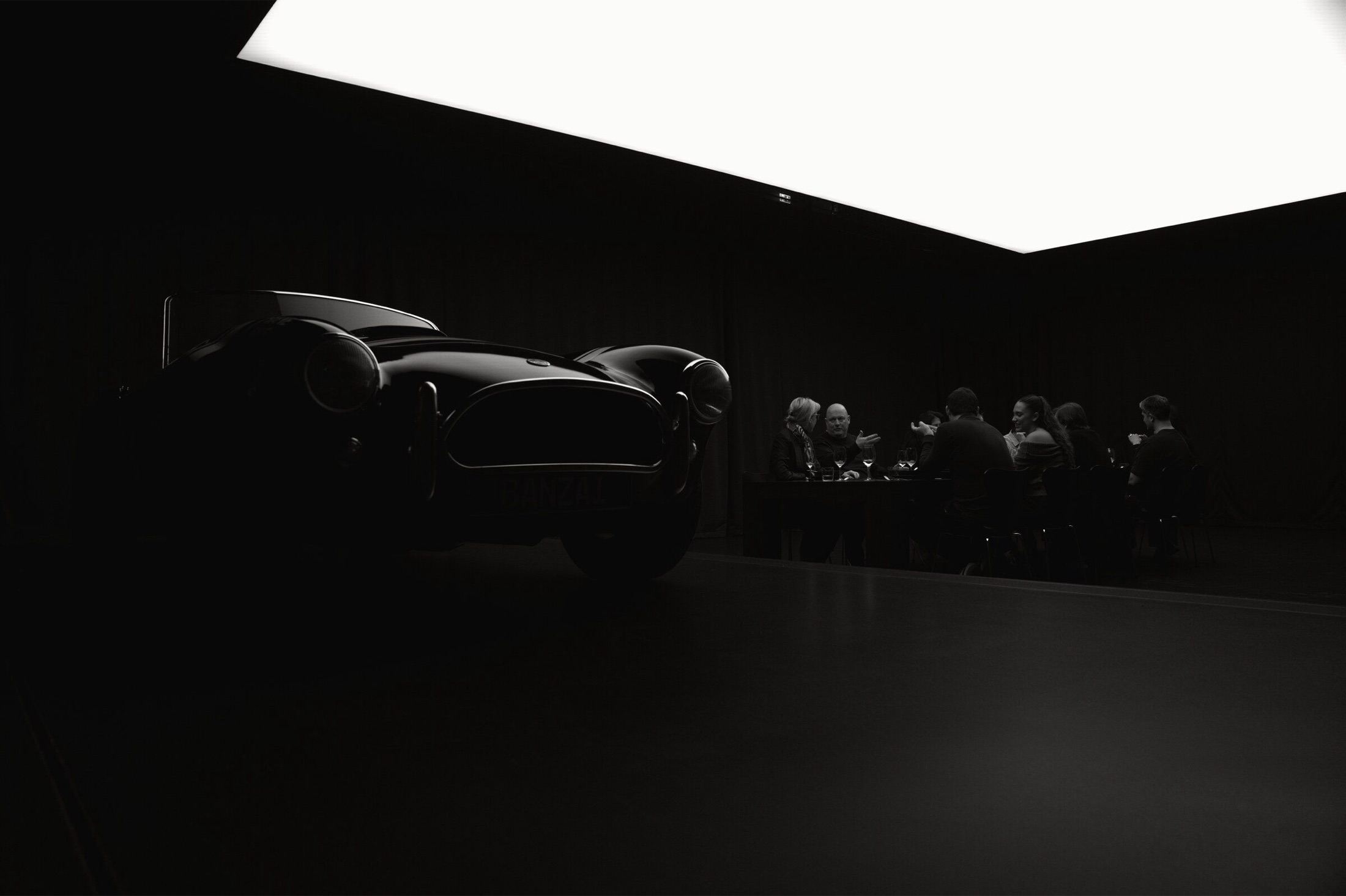 A collector car in silhouette while ten guests talk at a table in the background.
